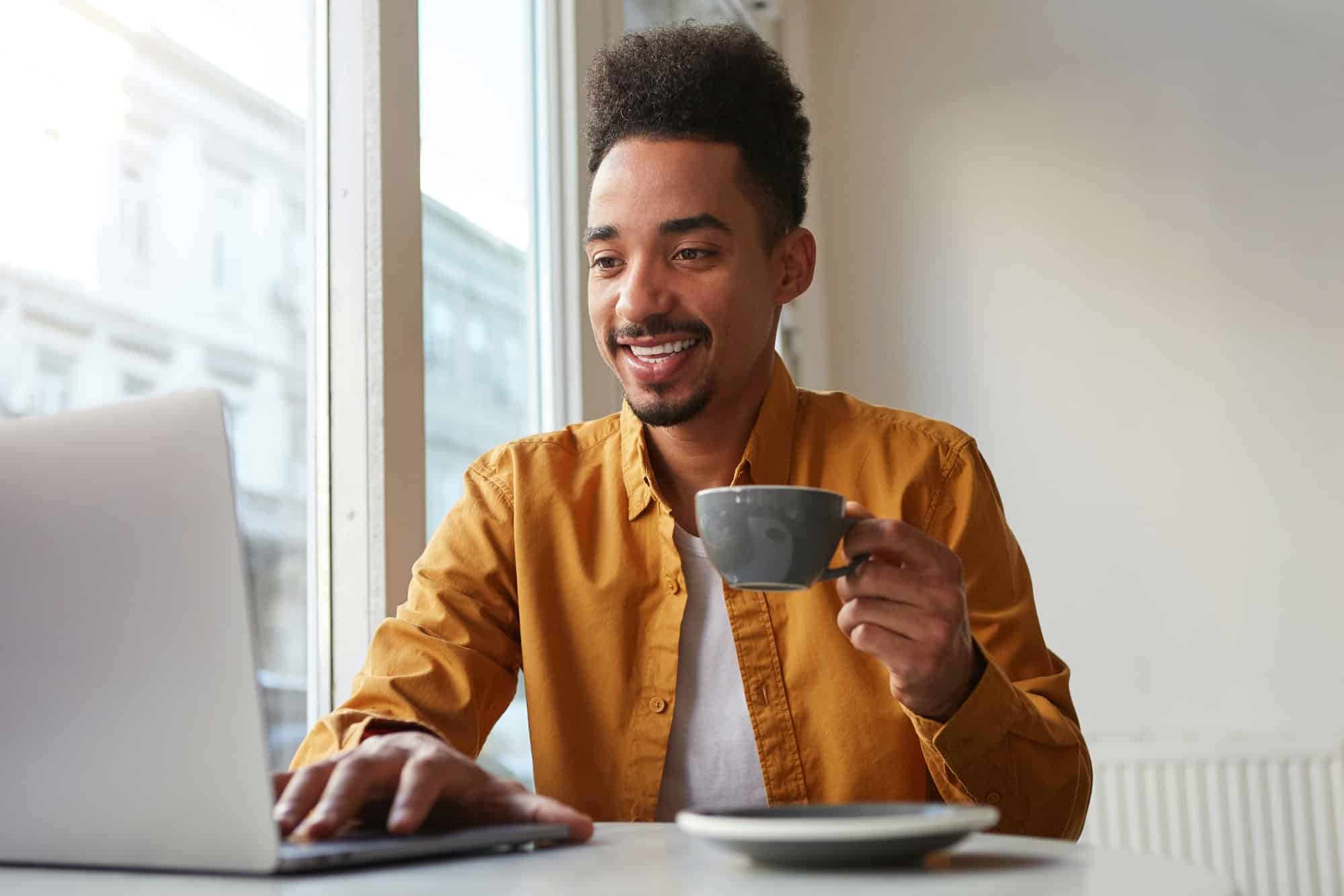Photo of glad African American attractive guy, sits in a cafe, creating at a laptop his new blog